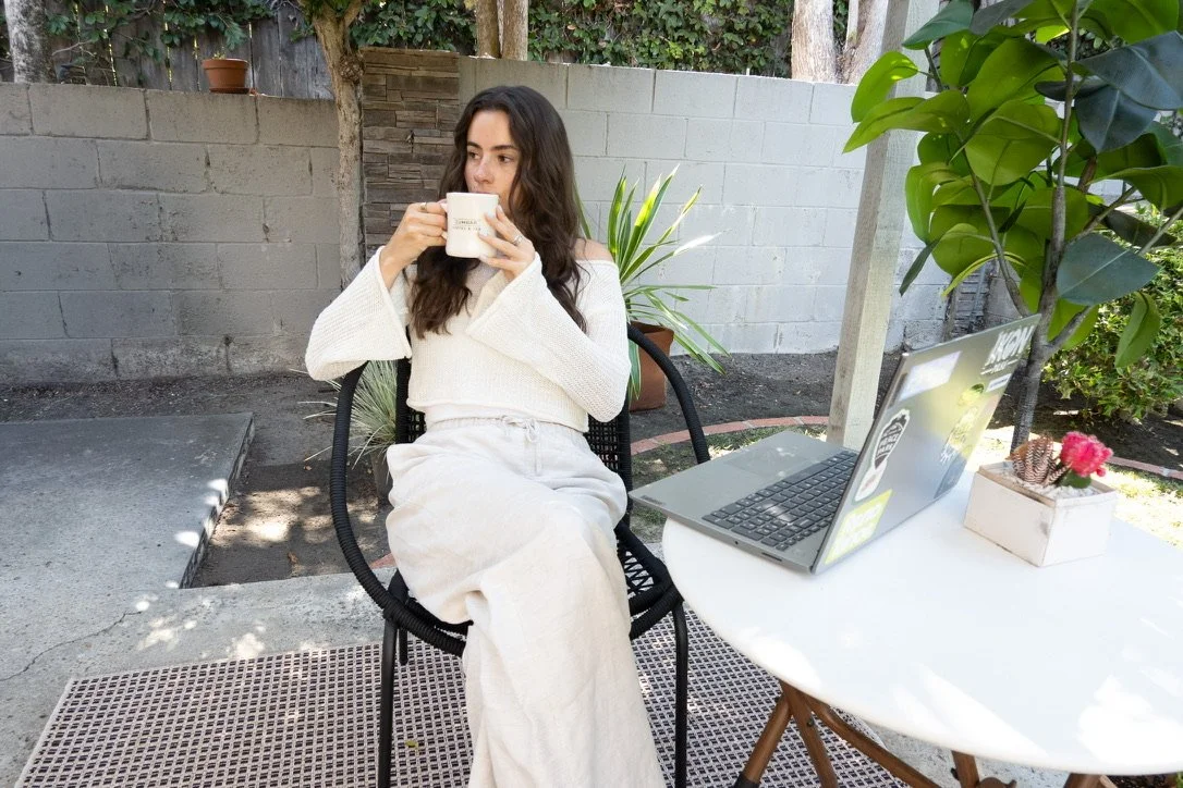 Woman enjoying coffee at an outdoor table with laptop, surrounded by plants and natural light.