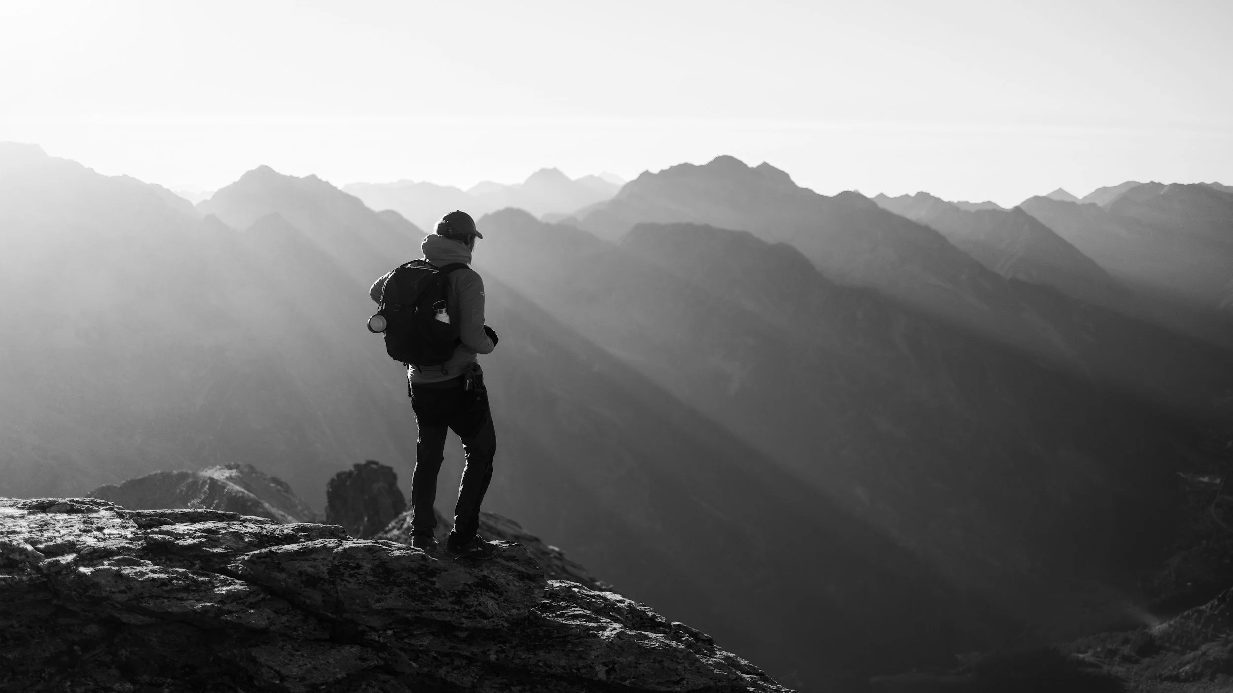 A lone hiker standing on a rocky ledge overlooking a mountain range in black and white.