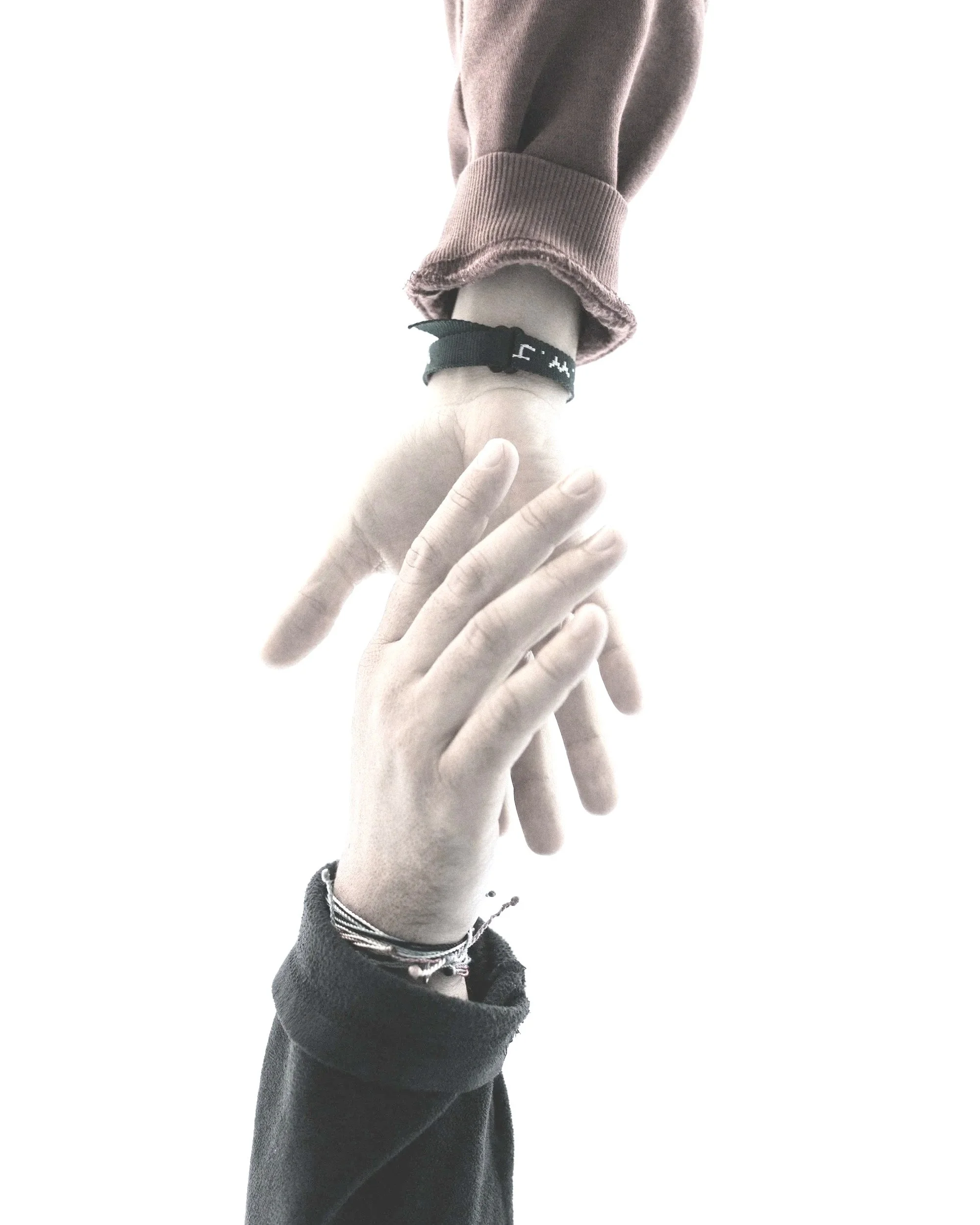 Close-up of a person's hand with fingers spread, wearing a black bracelet with white writing, shirt sleeve with frayed edges, and a sweatshirt sleeve with a ribbed cuff, against a bright white background.