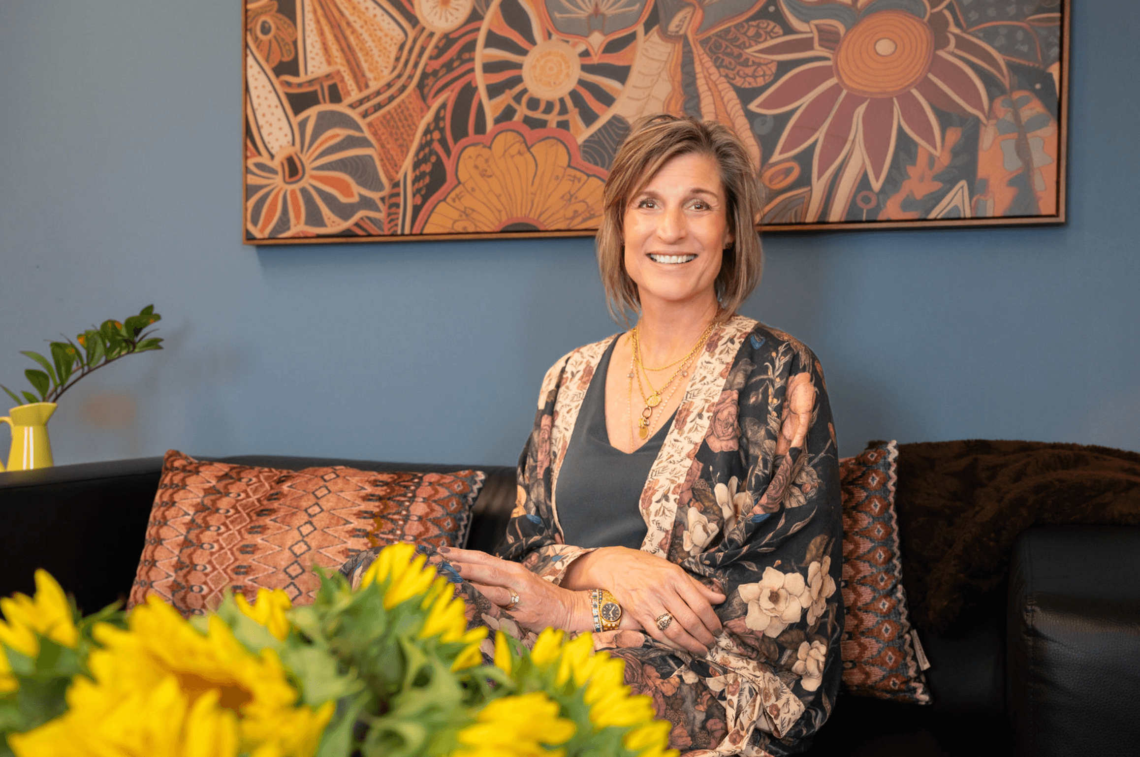 A smiling middle-aged woman with short, light brown hair sitting on a patterned brown sofa in front of a blue wall with a large, colorful abstract floral artwork. There is a vase of green leafy plant on the left, and yellow flowers in the foreground.