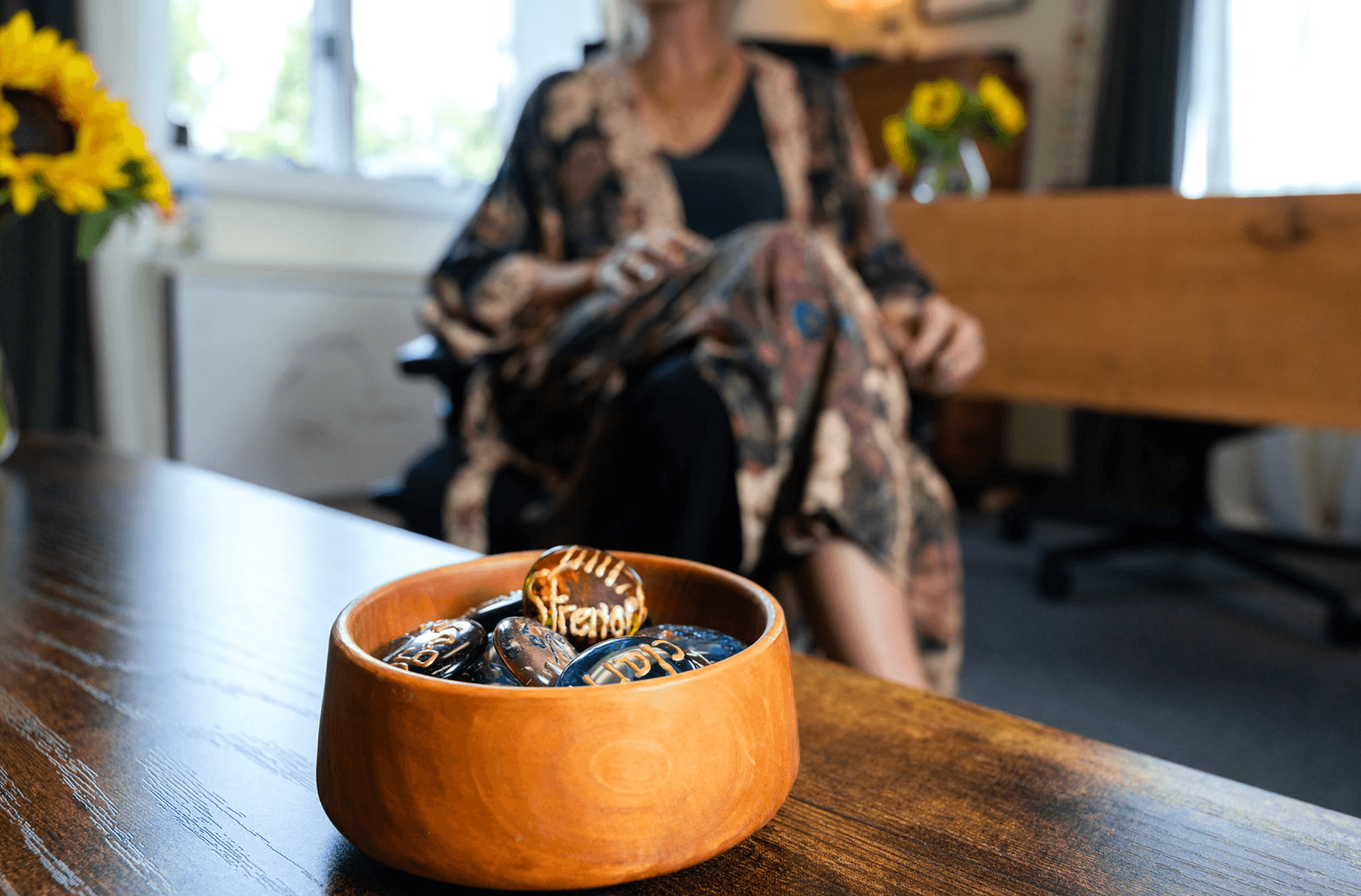 A bowl of chocolate-covered cookies on a wooden table with a woman sitting in the background, holding a phone, in a room with windows and a vase of yellow flowers.