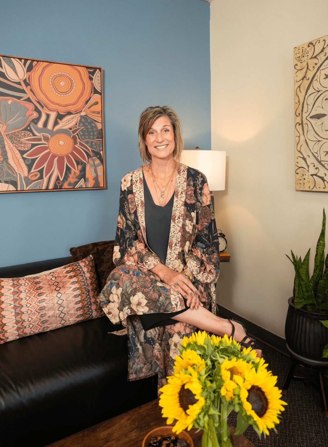 A woman sitting on a black leather sofa in a cozy, decorated room, smiling at the camera. She is wearing a floral robe over a black dress, with jewelry. There are colorful paintings on the wall behind her, a lamp, a green plant in a black pot, and a bouquet of sunflowers on a table in front.