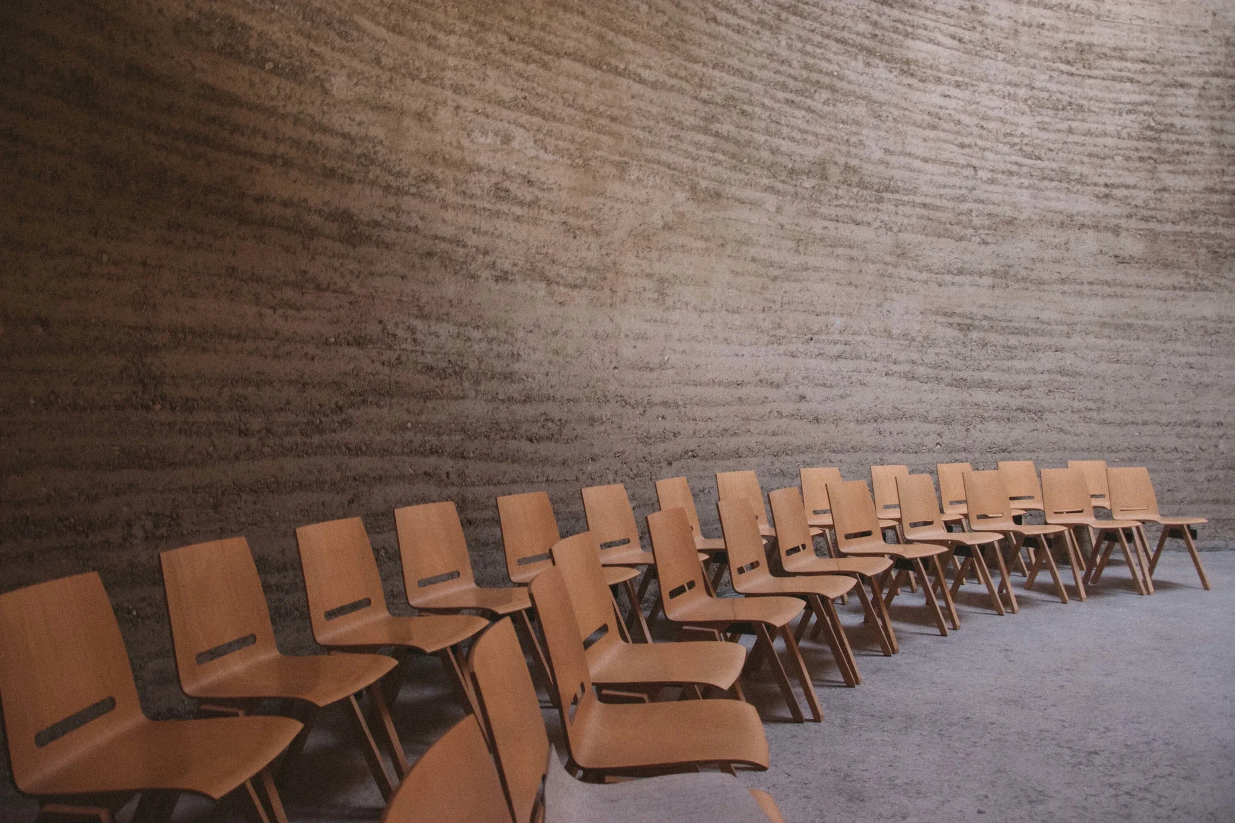 A line of wooden chairs arranged in an indoor space with a textured gray wall behind them.