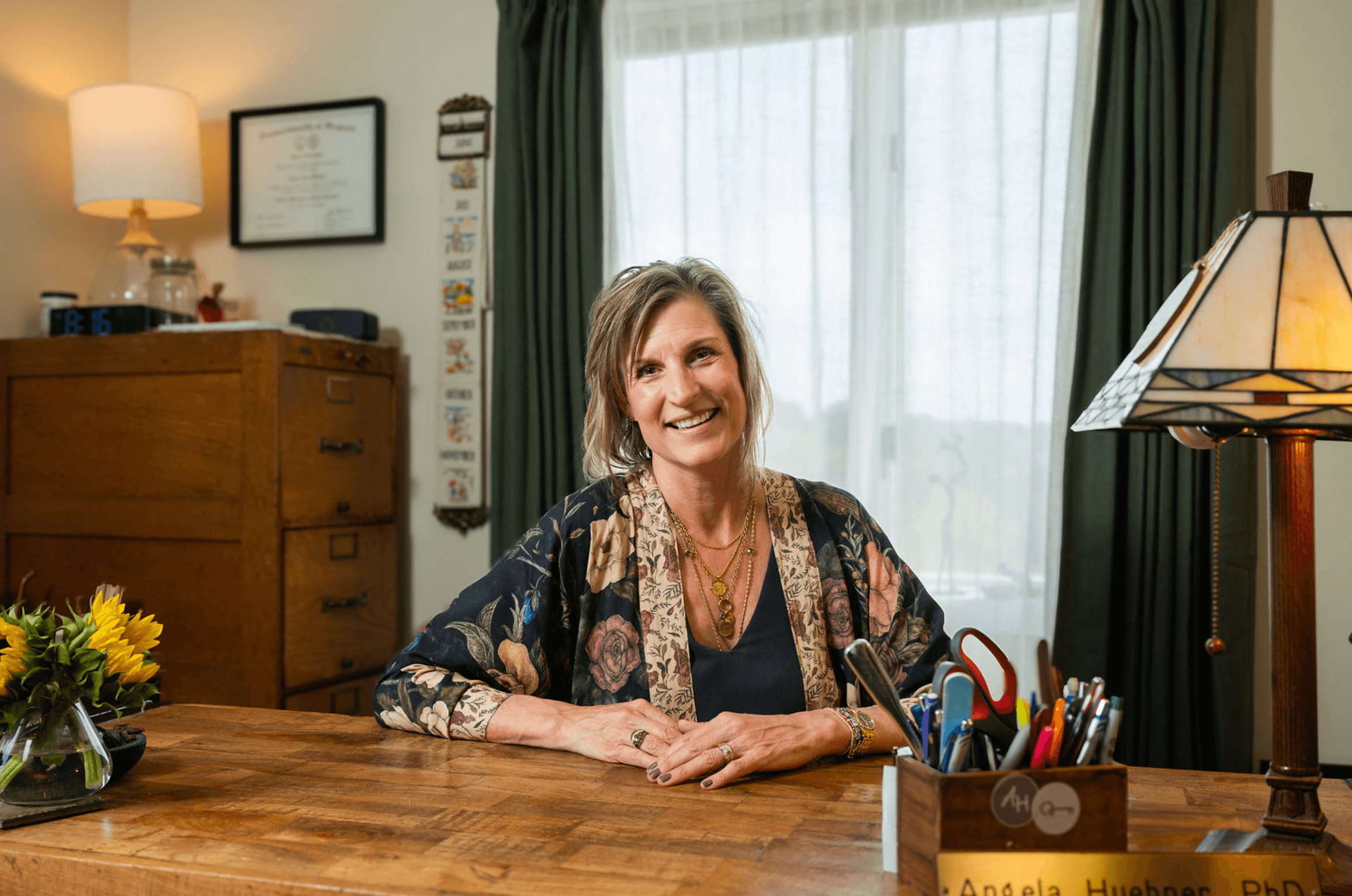 Angela Huebner, PhD, LMFT is sitting at a wooden desk in a cozy, well-lit room with green curtains and framed artwork on the wall, surrounded by office supplies and a vase of sunflowers.