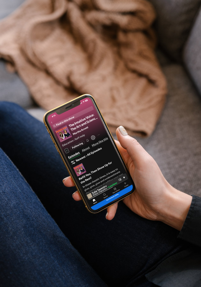 Person sitting on a gray couch, holding a smartphone with a podcast app open, displaying a podcast titled 'The Intuitive Voice: The Art and Science...'