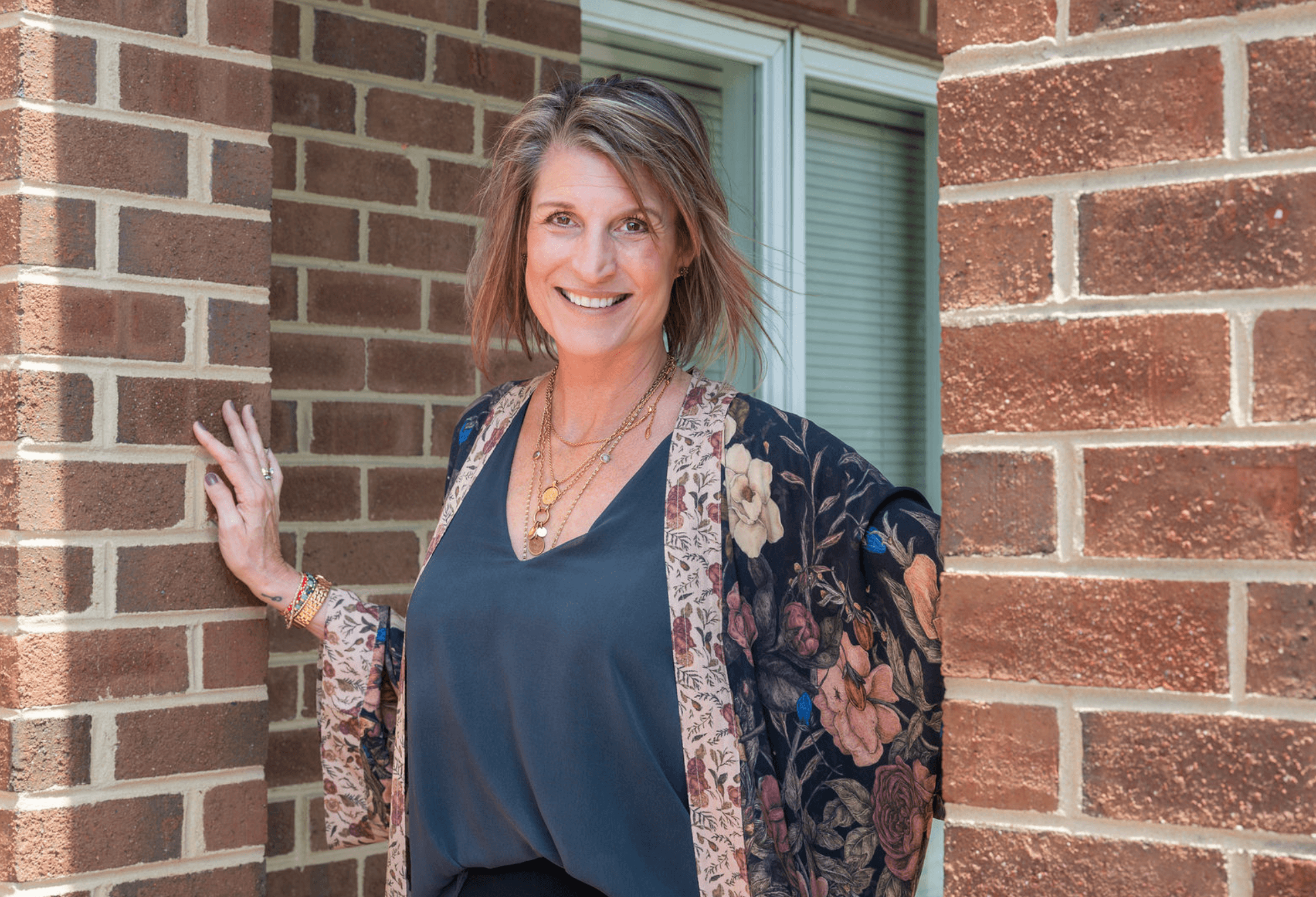 A smiling woman with shoulder-length brown hair standing outdoors near a brick wall, wearing a dark blue top, a floral jacket, and layered necklaces.