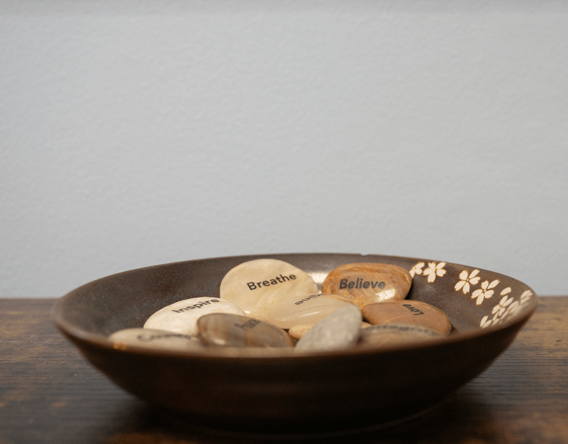 A round, dark-colored bowl containing smooth, flat stones with inspirational words such as 'Inspire,' 'Breathe,' and 'Believe' written on them, set on a wooden surface against a plain background.