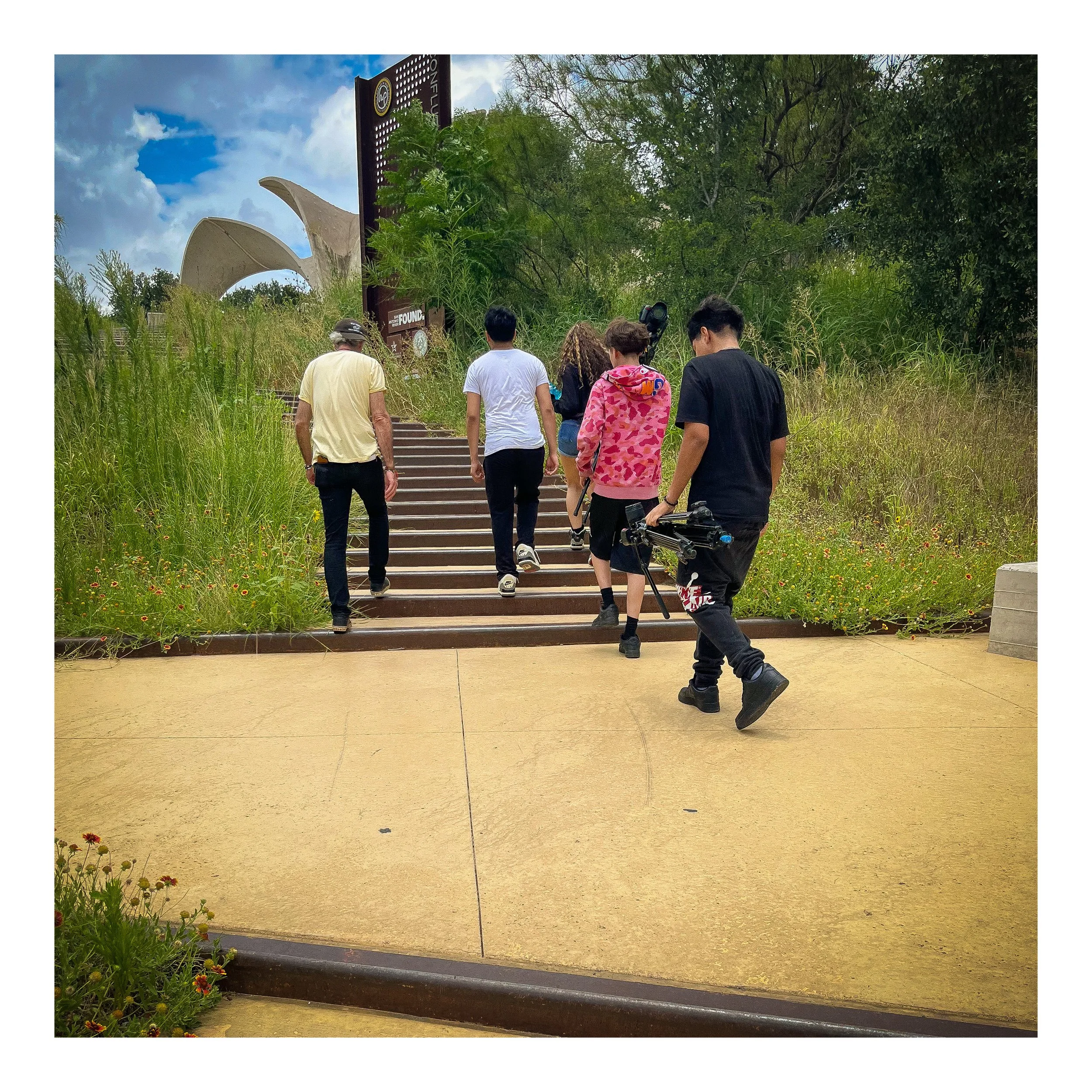 Group of people walking up outdoor stairs surrounded by greenery, with a large sculpture and a sign in the background under a partly cloudy sky.