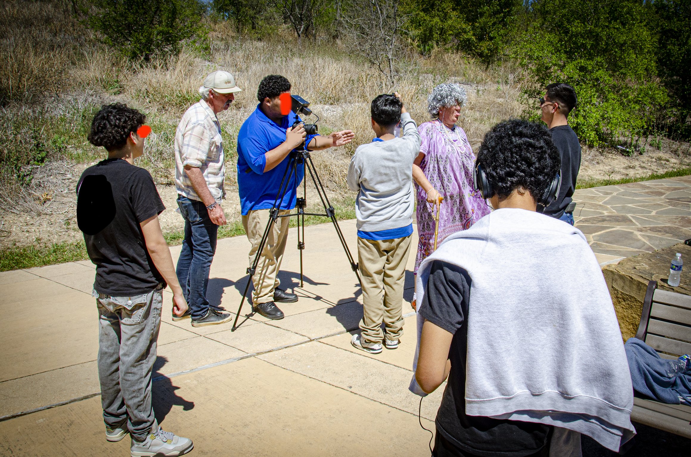 Group of people outdoors, with some wearing headphones, interacting, and a person operating a camera on a tripod.