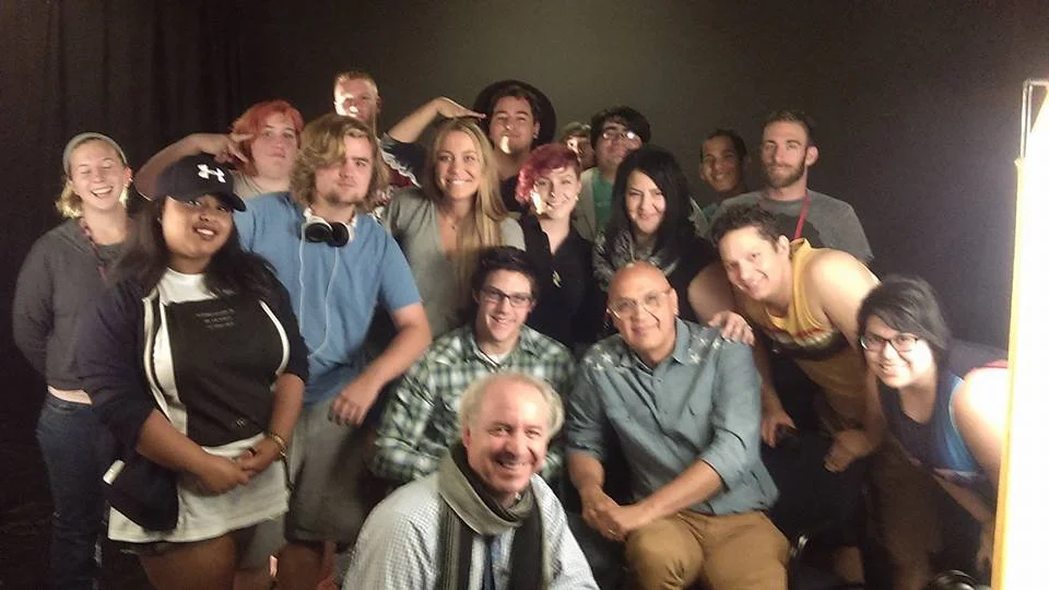 Group of 15 people, mostly young adults, smiling and posing together in a dimly lit room, some making playful gestures.