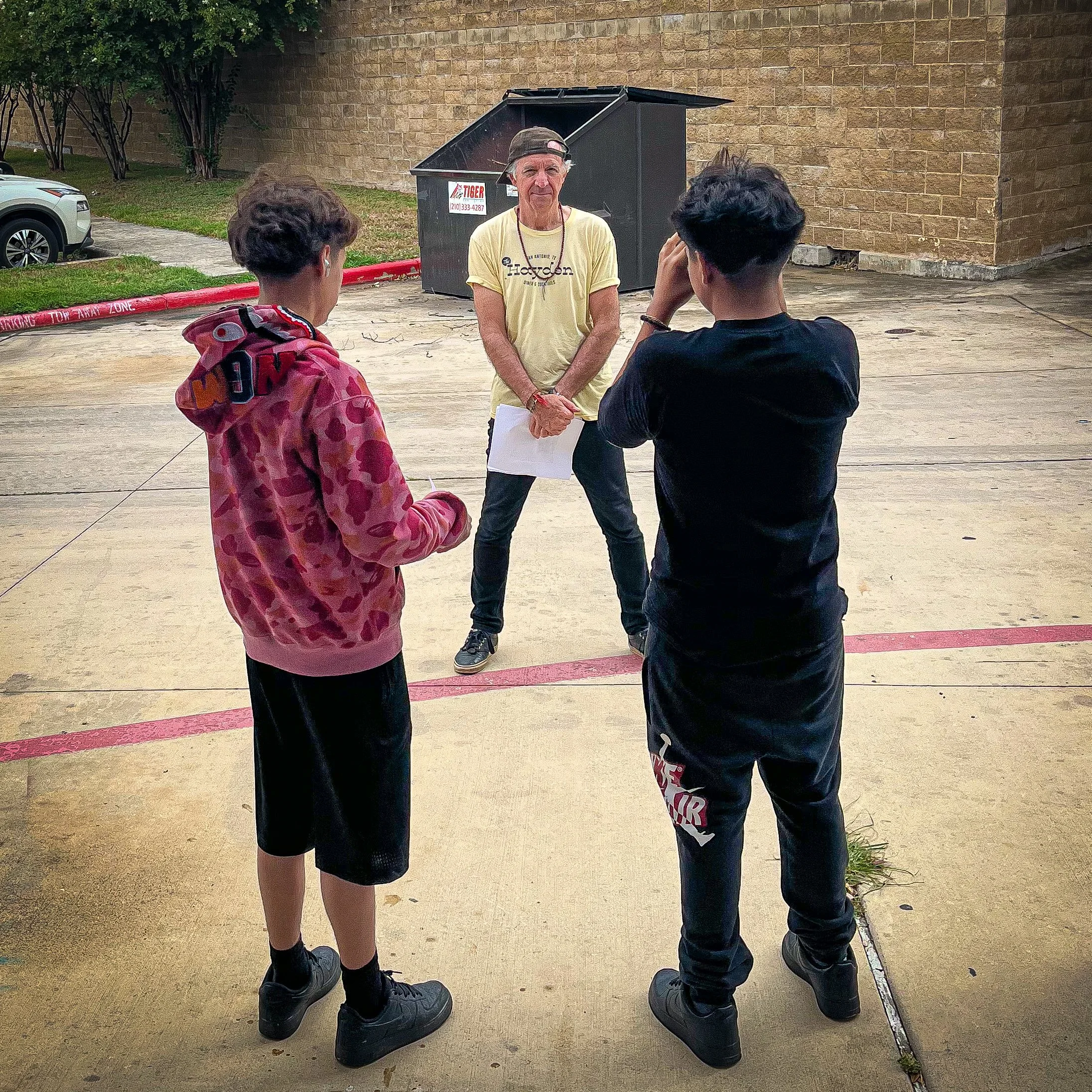 A man with a backwards cap and yellow shirt standing outdoors, talking to two children, one wearing a pink hoodie and the other in black. The children are listening, and the setting appears to be a parking lot with a brick wall, cars, and a dumpster in the background.