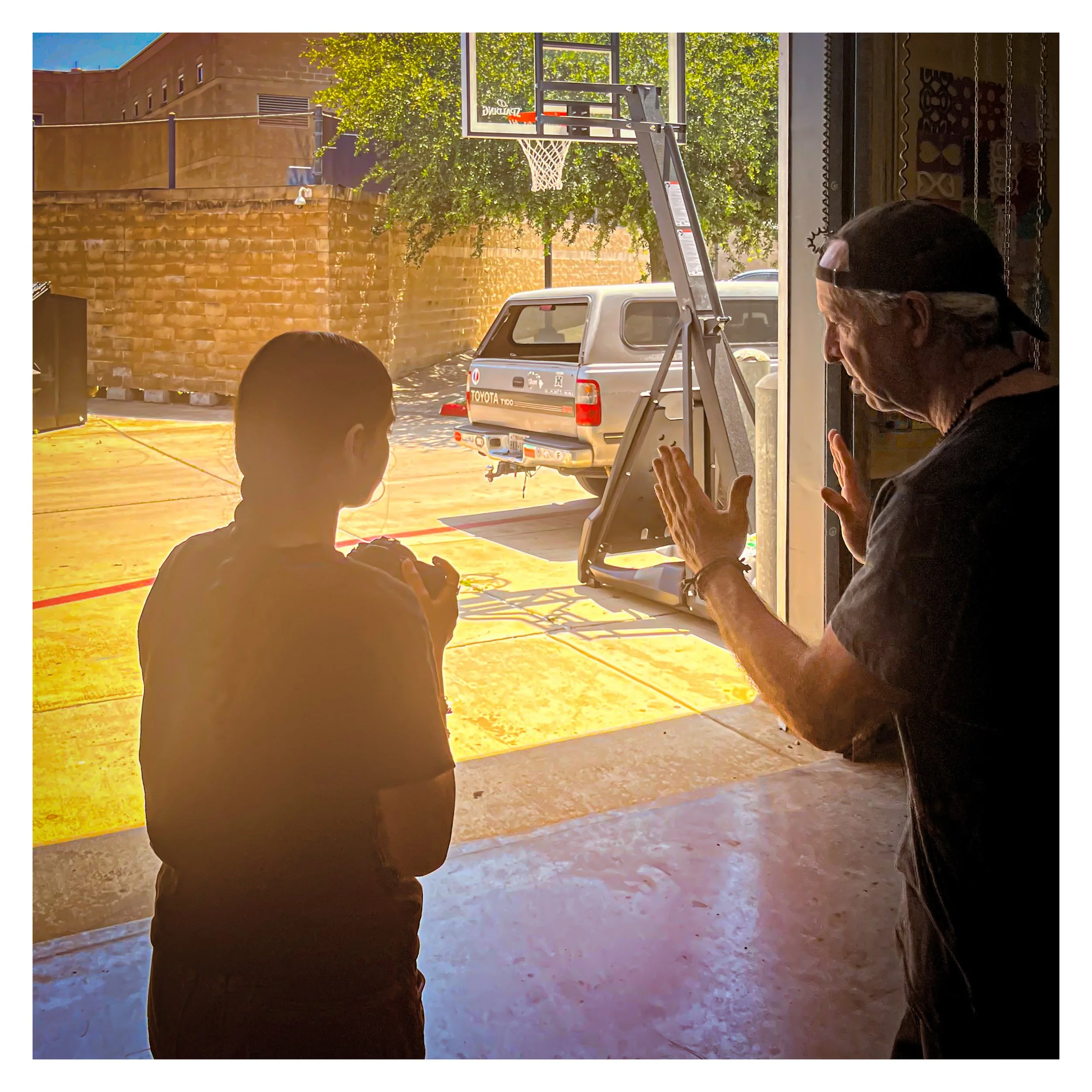 People inside a building looking out through large open garage doors at a parking lot with a pickup truck, basketball hoop, brick wall, trees, and blue sky outside.