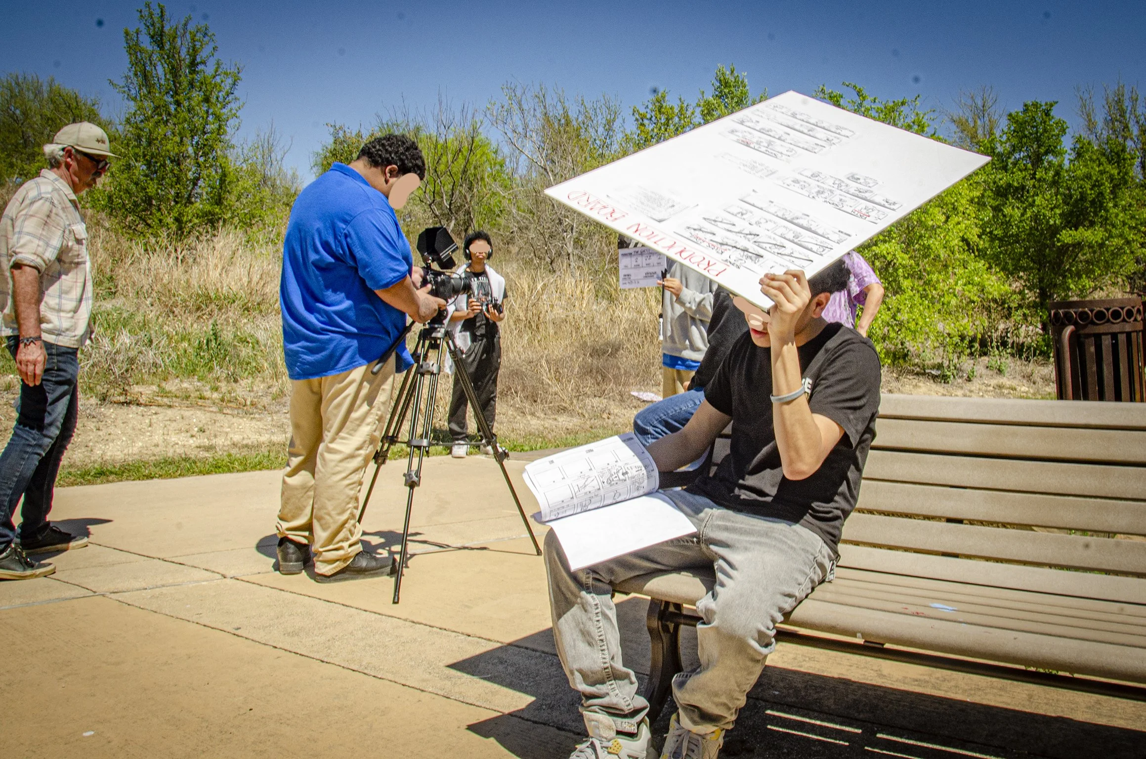 Group of people outdoors, one person sitting on a bench holding a large map, others standing nearby, some with cameras, in a natural setting with trees and a clear blue sky.