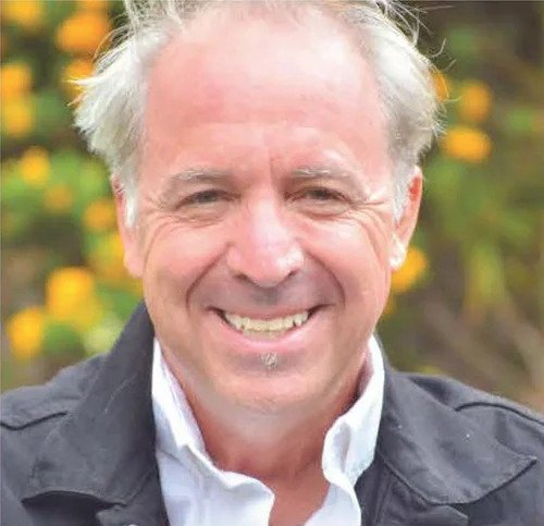 A smiling middle-aged man with light-colored hair and a white shirt, outdoors with flowers in background.