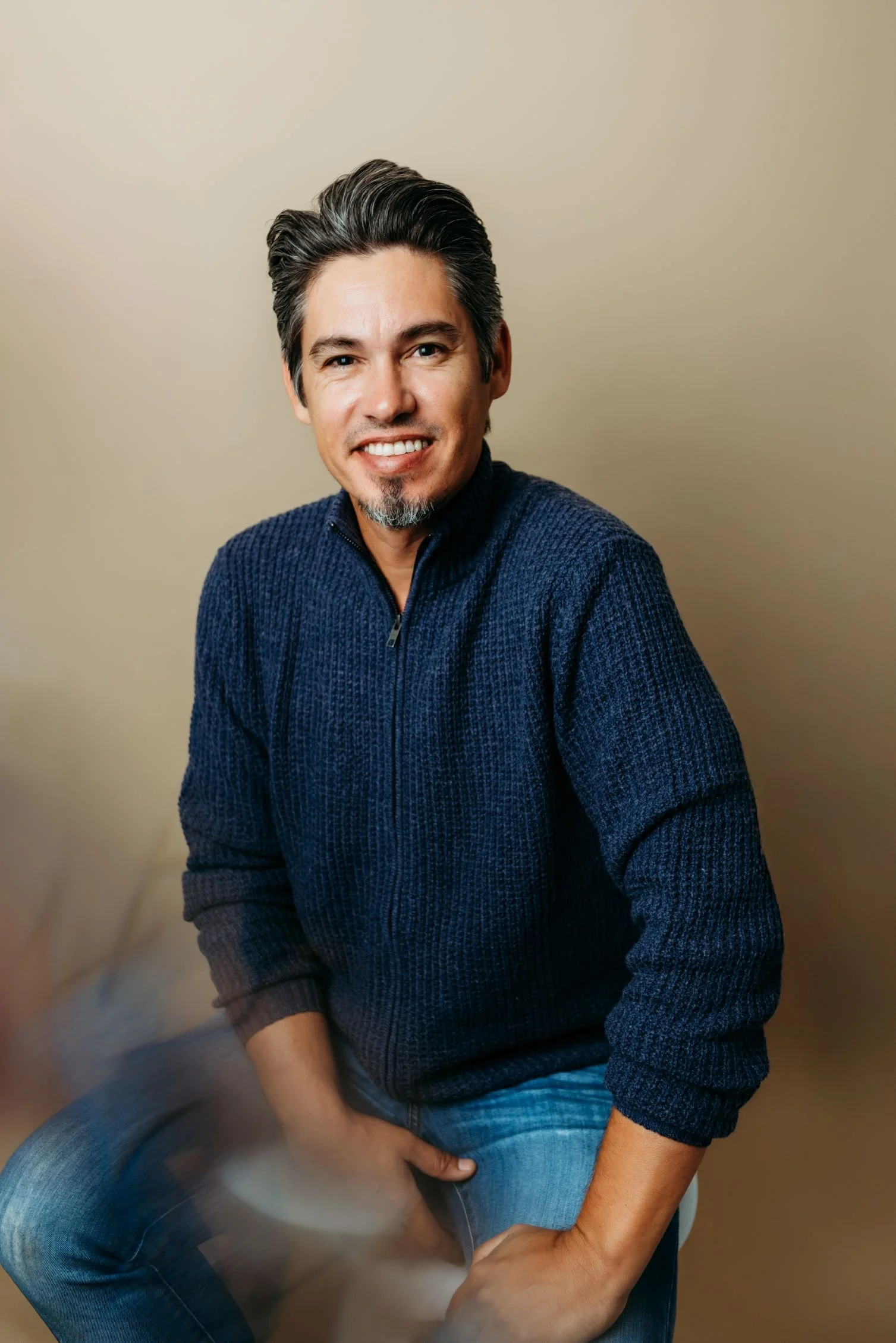 A man with dark hair, a beard, wearing a dark blue sweater and blue jeans, sitting on a stool against a plain beige background.