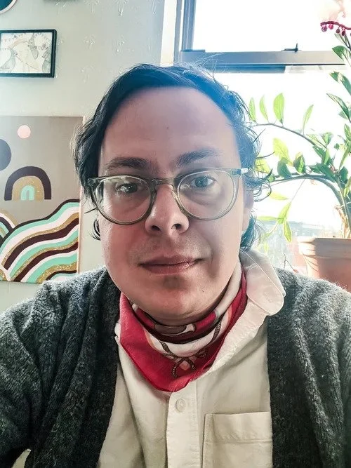 Person with glasses and dark hair, wearing a white shirt, red bandana, and dark cardigan, sitting near a window with plants and artwork in the background.