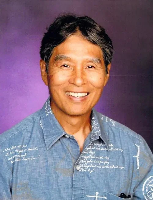 Portrait of a smiling middle-aged Asian man with short black hair, wearing a blue denim shirt, against a purple background.
