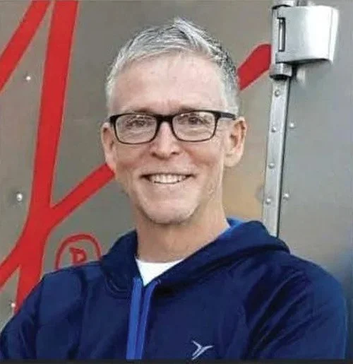 A smiling man with short gray hair, glasses, and a navy blue jacket standing outdoors.