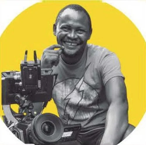 A man with dark skin, smiling and sitting next to a professional camera on a tripod, against a yellow circular background.