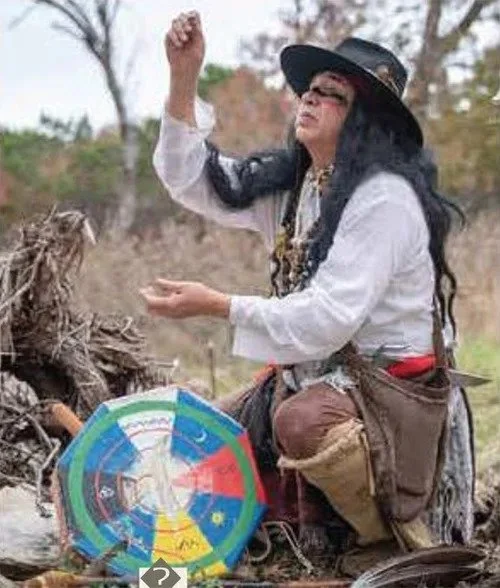 A woman dressed as a witch in a Halloween costume, with a black hat and long black hair, sitting outdoors. She is holding a colorful spinning wheel for a game or fortune-telling, and appears to be talking or gesturing with her right hand.