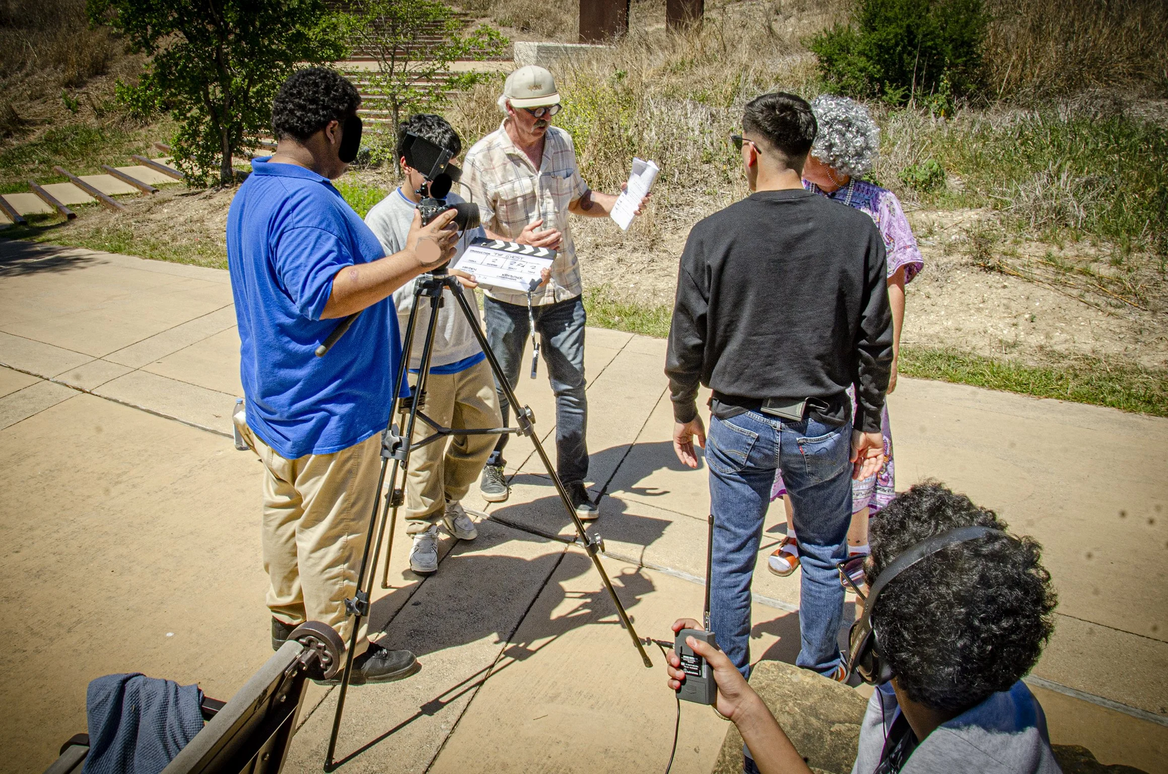 A group of people, including a man with a clipboard, a person with a clapperboard, and a camera operator, filming an older woman with curly gray hair outdoors on a sunny day with dry grass and bushes in the background.