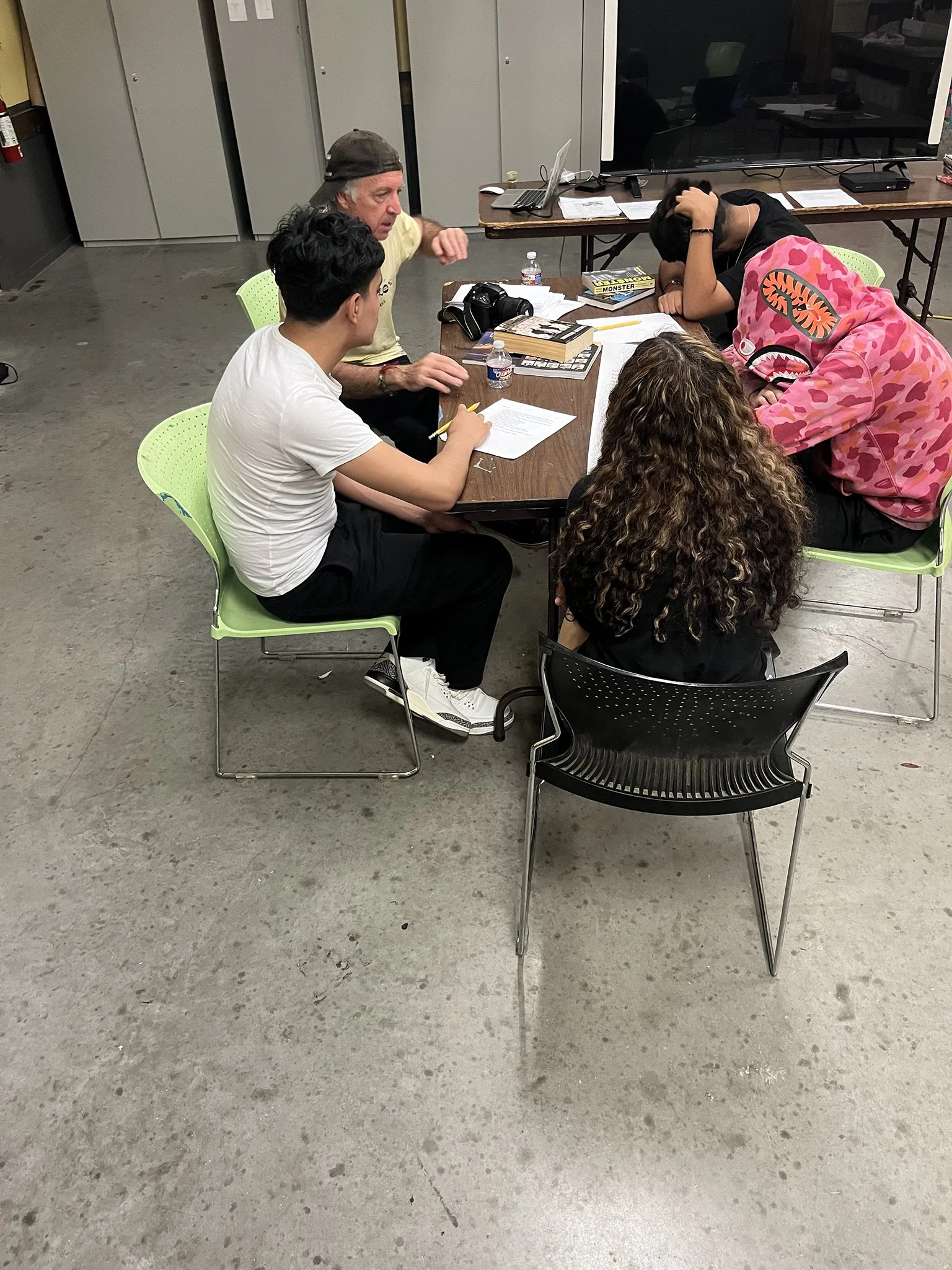 Five people sitting around a table with papers, books, and water bottles, engaged in conversation in an industrial-style room with a concrete floor and storage cabinets.