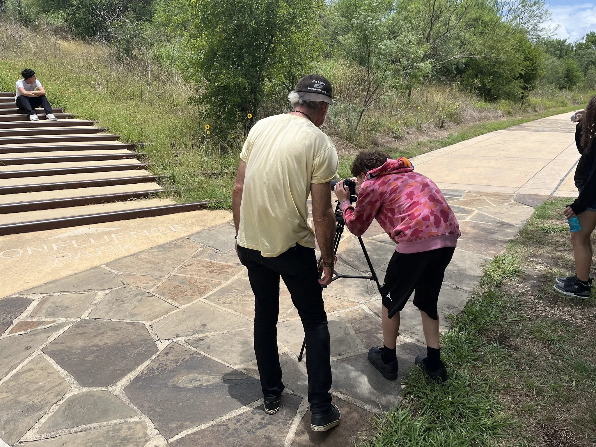 Four people are outdoors on a paved area and grassy hillside. One young man sits on stairs to the left, while a man and a woman with a camera stand near the middle. Another person stands on the right, partially out of the frame. Trees and a cloudy sky are in the background.