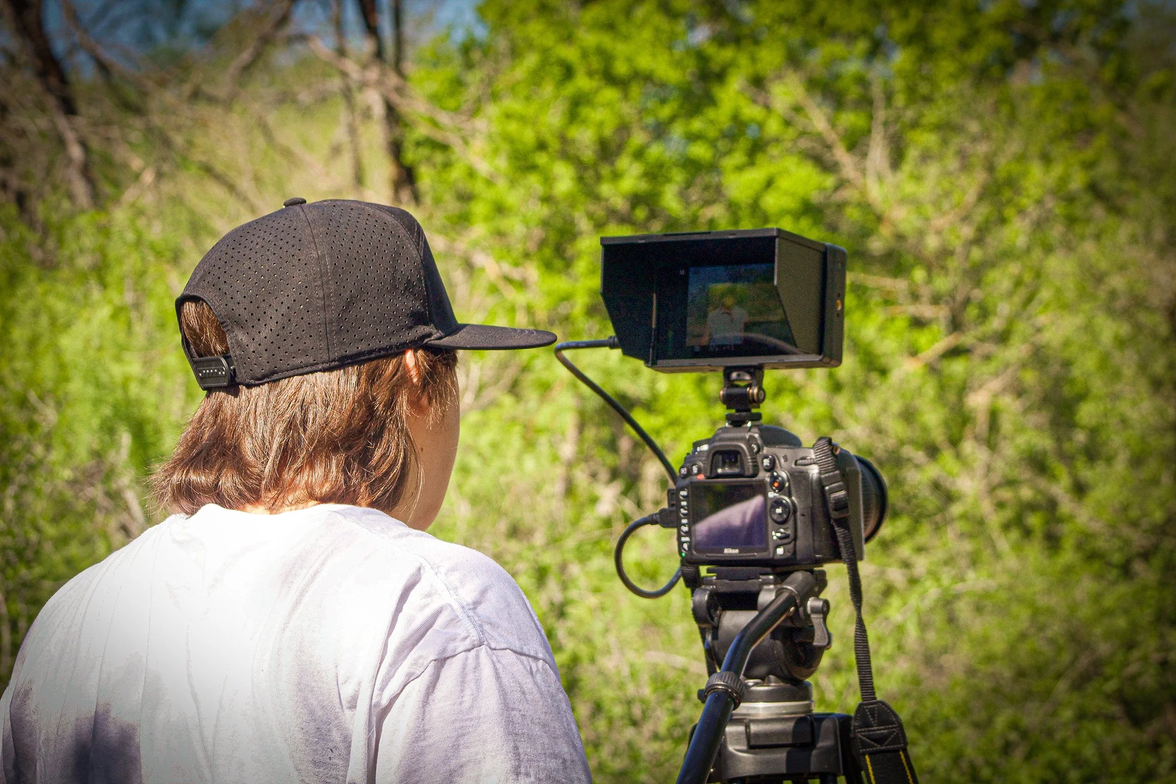 Person with long brown hair wearing a black baseball cap using a camera on a tripod outdoors in a green forested area.