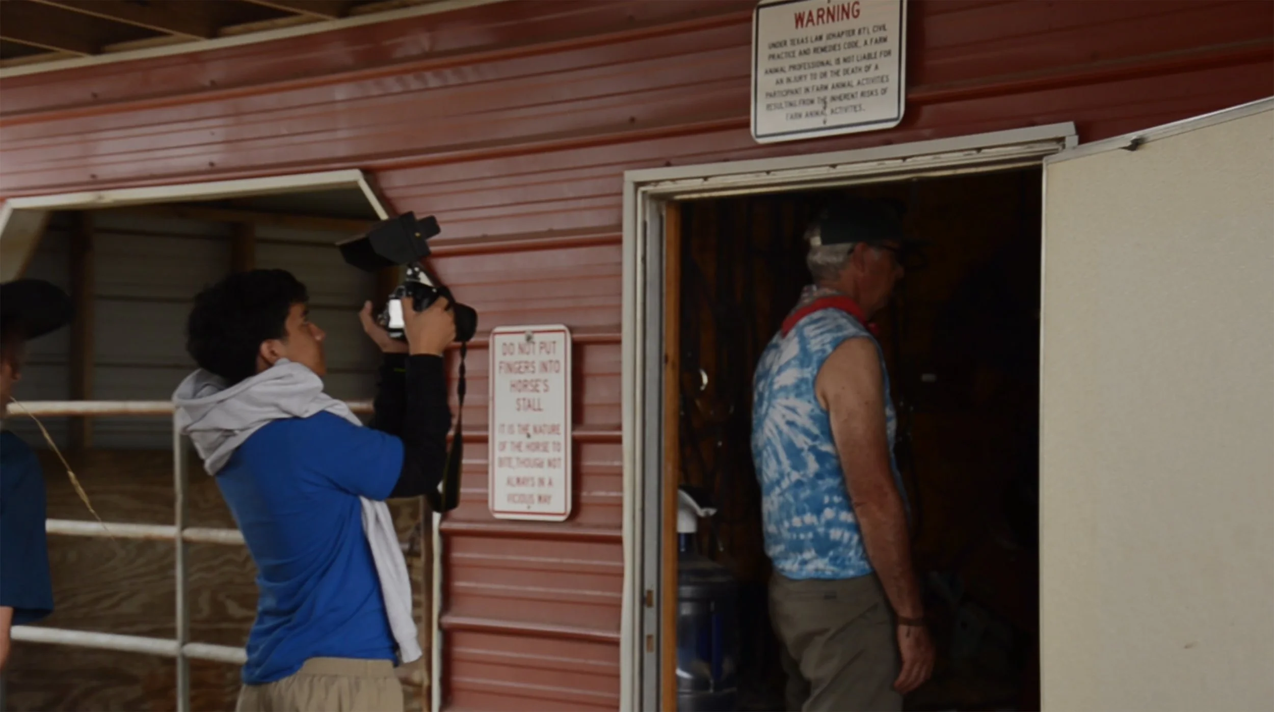 A young man with a camera taking a photo of an older man inside a small building, with a woman standing nearby. The building has red walls and multiple signs, including a warning sign and a notice about not putting fingers into horse stalls.