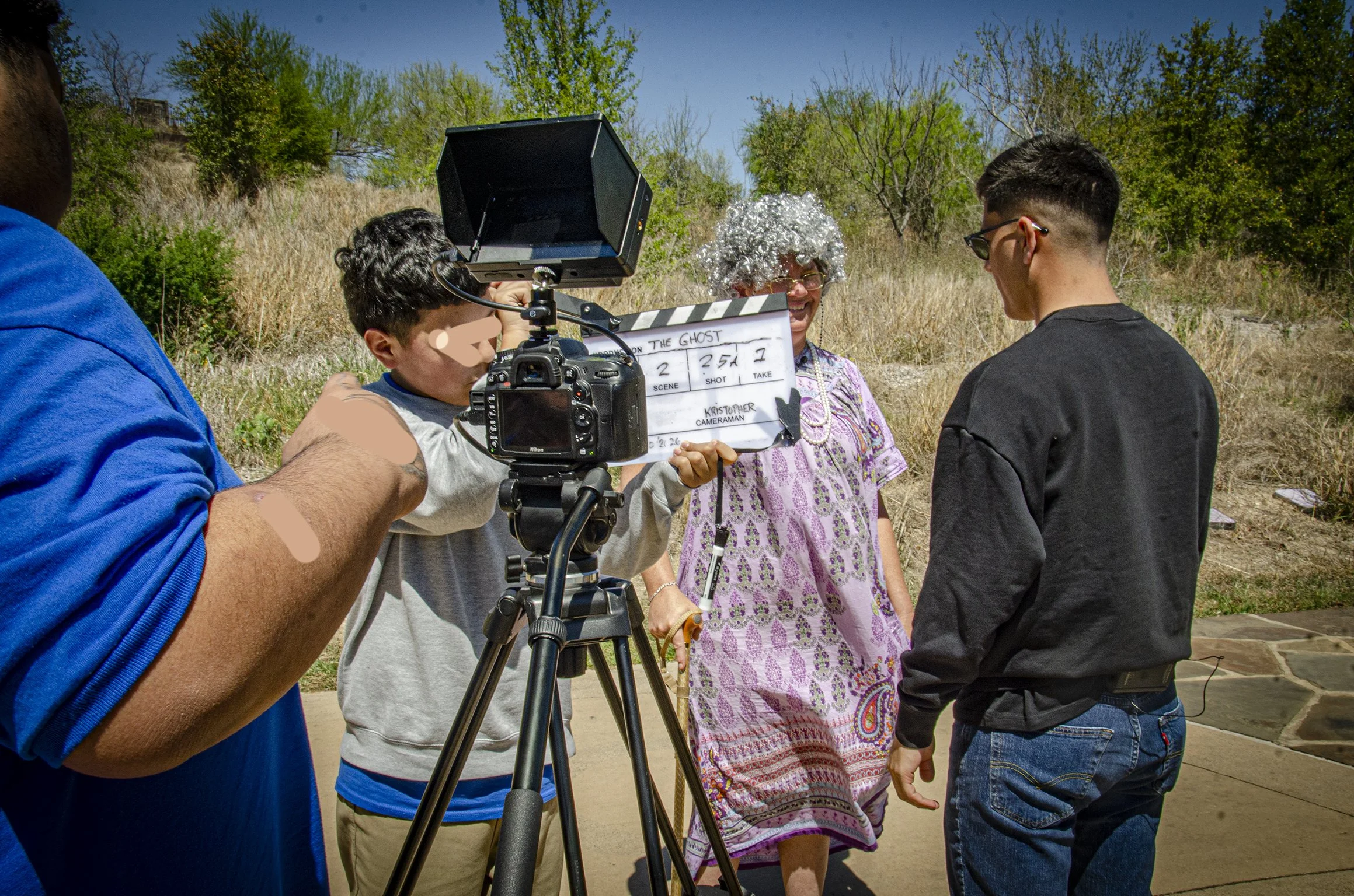A film crew is filming a woman dressed as a ghost outdoors on a sunny day, with trees and bushes in the background.