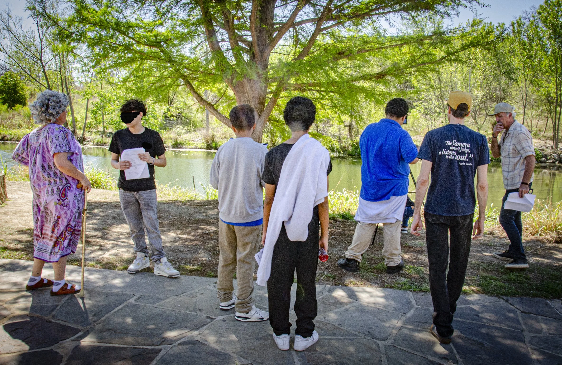 A group of eight people, including children and adults, standing outdoors by a body of water under a large green tree. One person is wearing a purple patterned dress with a wig, another is holding a paper, and others are looking at their phones or talking. The scene appears to be a casual gathering or outdoor activity in a park.
