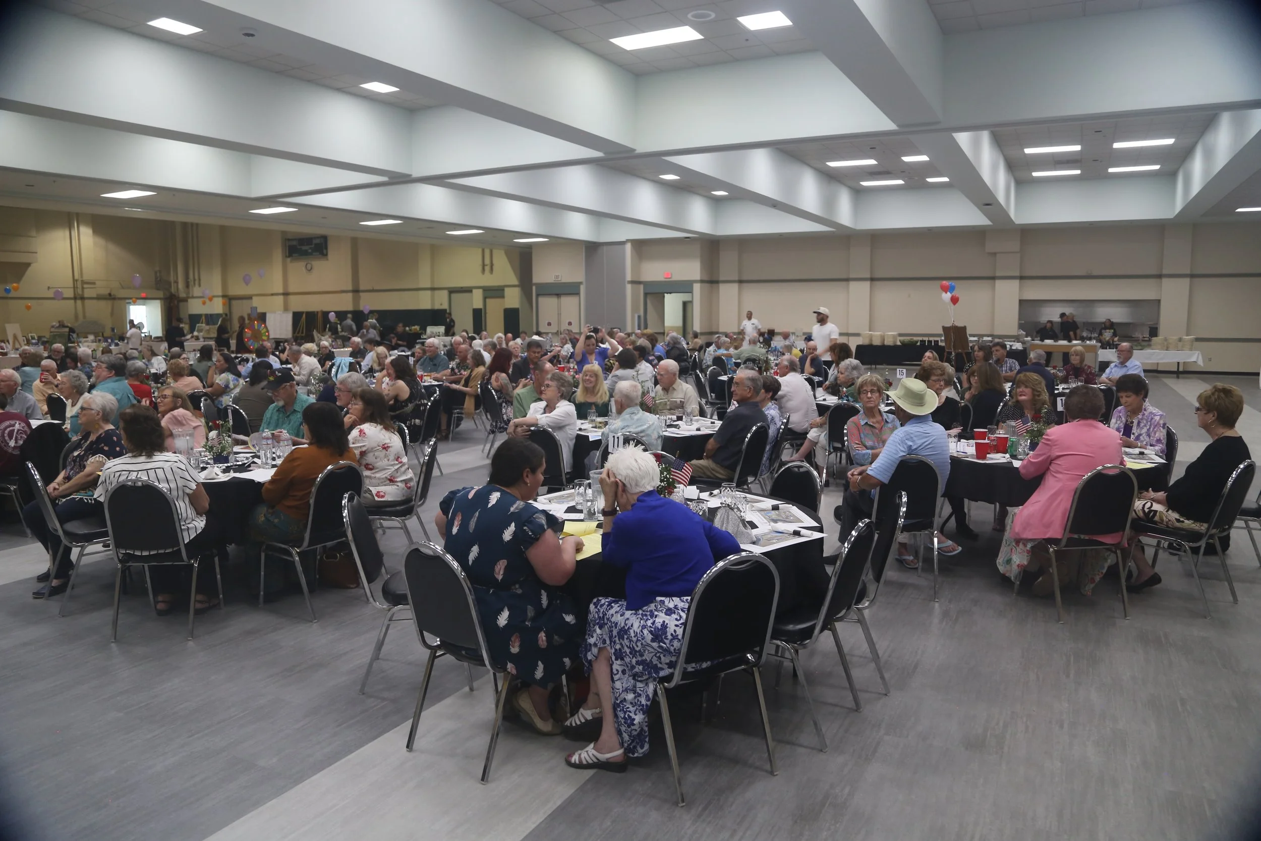 Large indoor event with many people sitting at round tables decorated with patriotic items like small American flags, in a spacious hall with high ceiling and bright lighting.