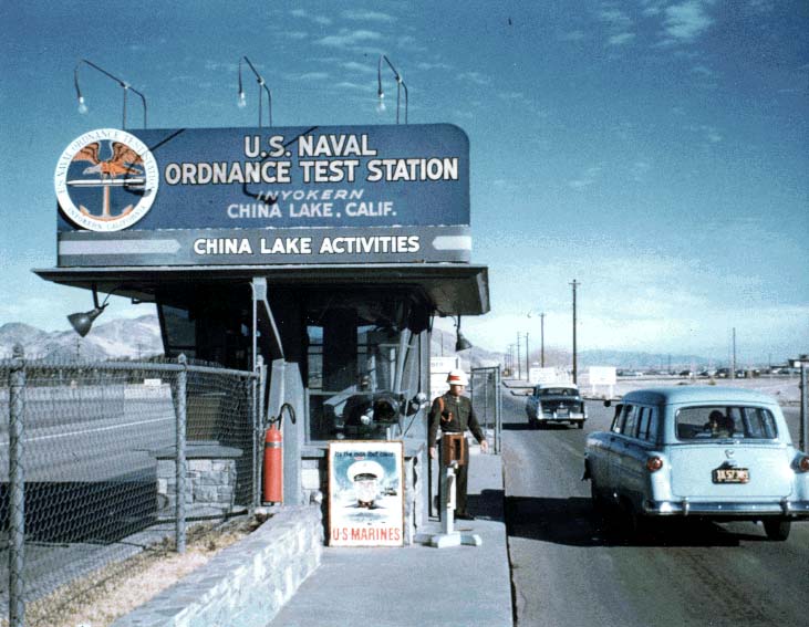 U.S. Naval Ordnance Test Station sign at China Lake, California main gate, with a man in uniform standing near a small guard booth, and 1940's vintage cars parked on the street.