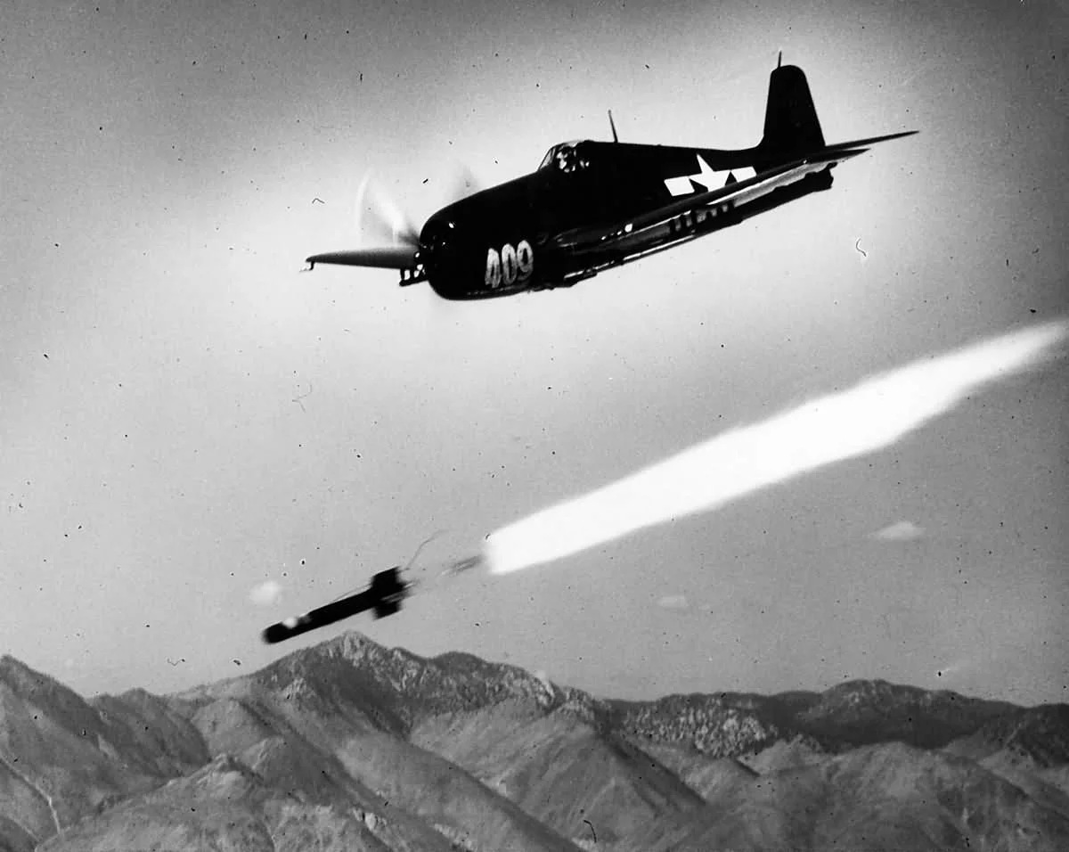 Black and white photo of a fighter plane in mid-air releasing a missile, with mountainous terrain below.
