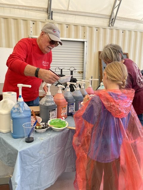 Older man with sunglasses and a red shirt applying colored paint or dye to a young girl in a cape at a STEAM art class
