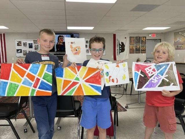 Three children in a classroom holding up colorful abstract paintings and drawings they made.