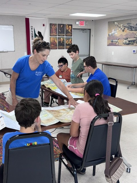 Teacher instructing a group of children seated at tables with maps in a classroom decorated with posters and aircraft murals.