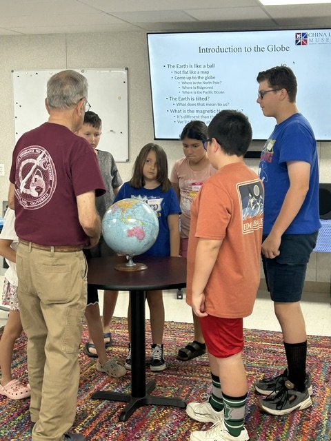 A man teaching a group of five children about the globe in a classroom. There is a large screen in the background displaying information about the Earth and the Globe. The children are gathered around a table with a globe on it, paying attention to the man.