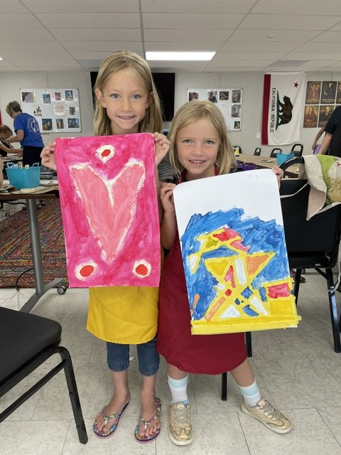 Two young girls holding colorful abstract paintings inside an art classroom.