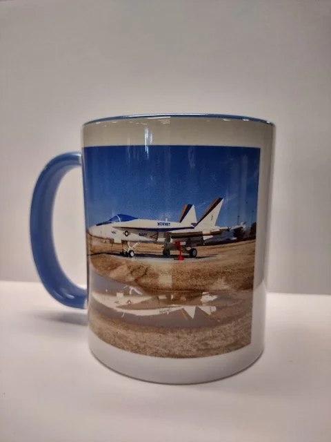 A white coffee mug with a blue handle featuring a photo of a fighter jet at the China Lake Museum.