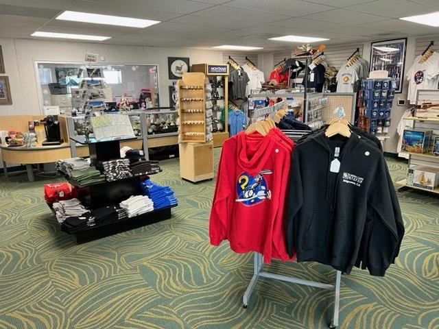 Inside a retail store with various clothing items, hats, and souvenirs displayed on shelves, racks, and tables.