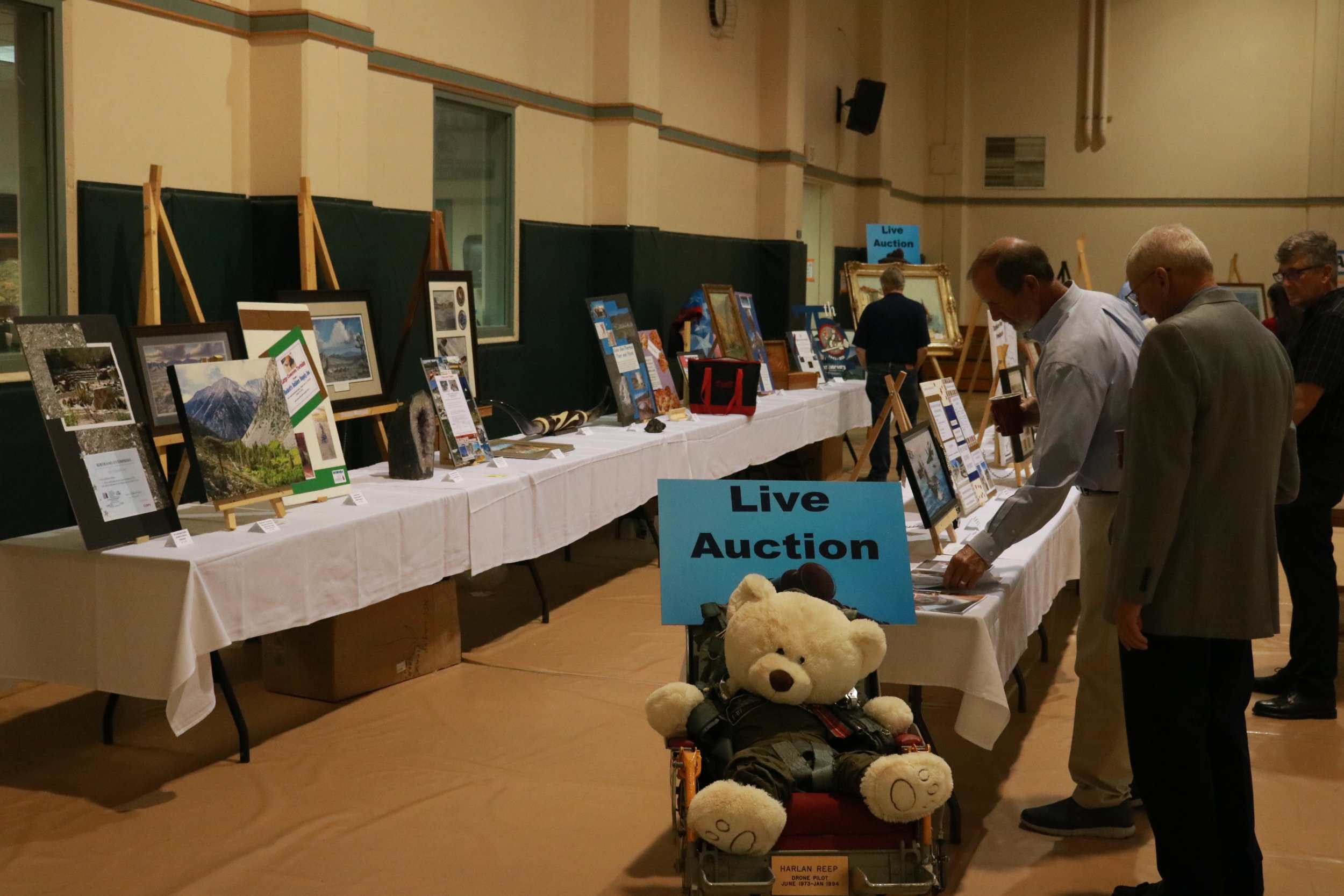 Indoor auction event with artwork and framed pictures on display tables, and people browsing items, including a stuffed teddy bear at the front.