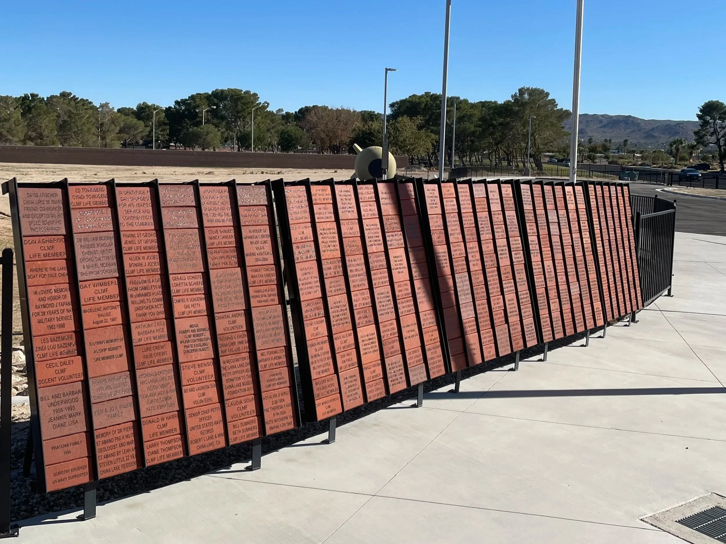 A memorial wall with numerous red bricks, each inscribed with names and messages, situated outdoors against a backdrop of trees, mountains, and a clear blue sky.