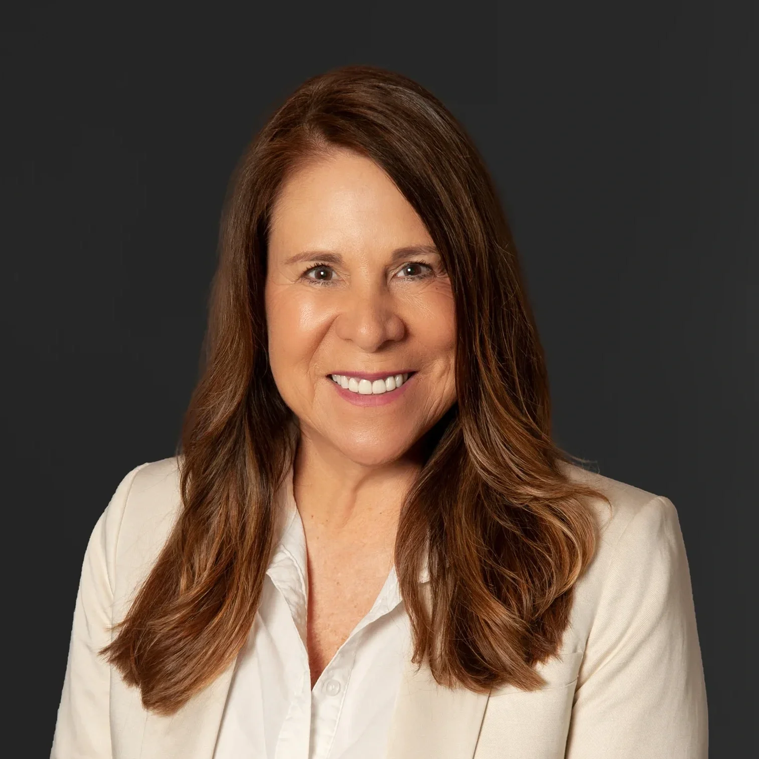 A woman with long, wavy, reddish-brown hair wearing a white blazer and shirt, smiling and looking at the camera against a dark background.