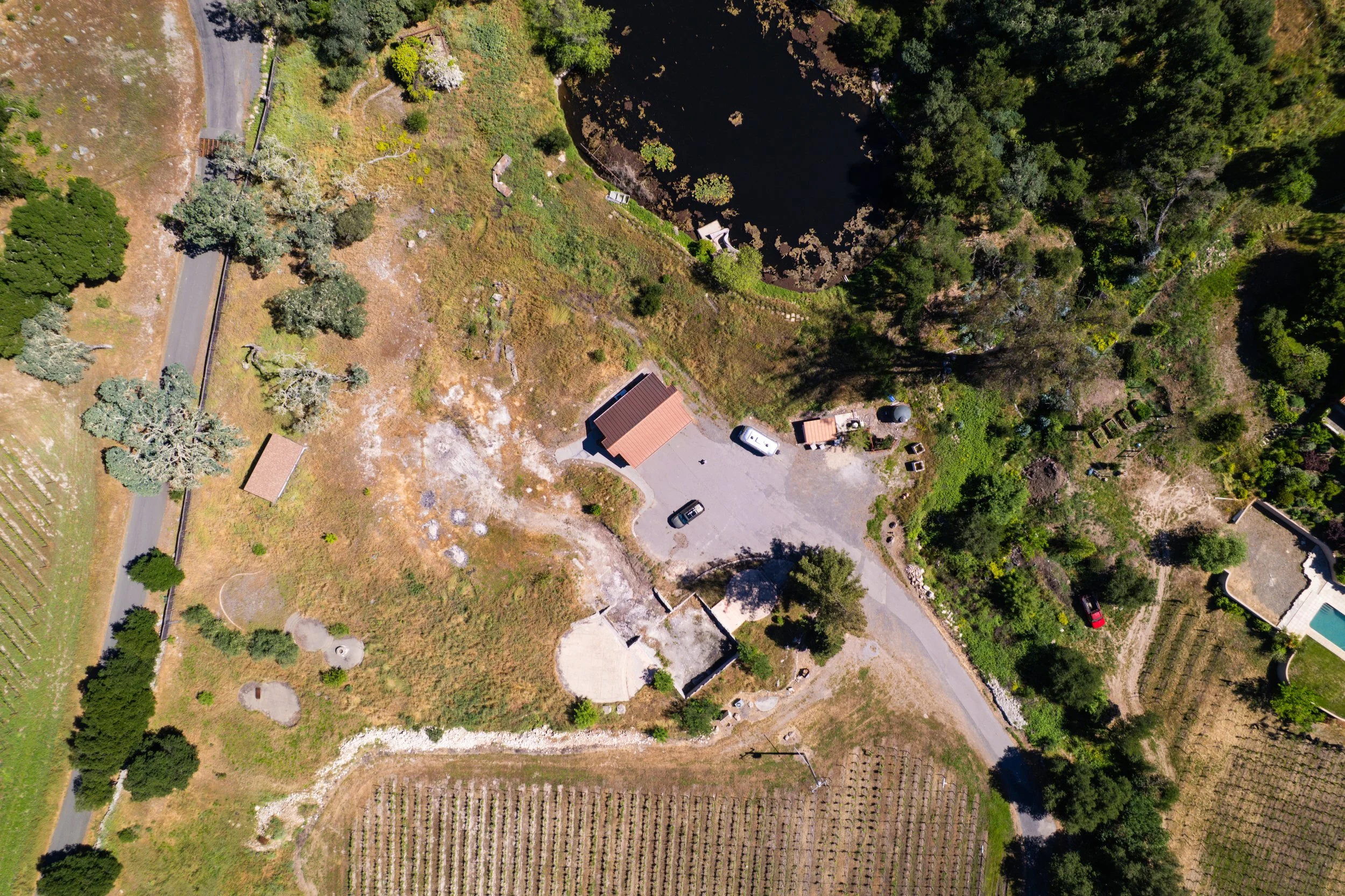 An aerial view of a rural area showing a pond surrounded by trees, a building, a parking lot with cars, and farmland.