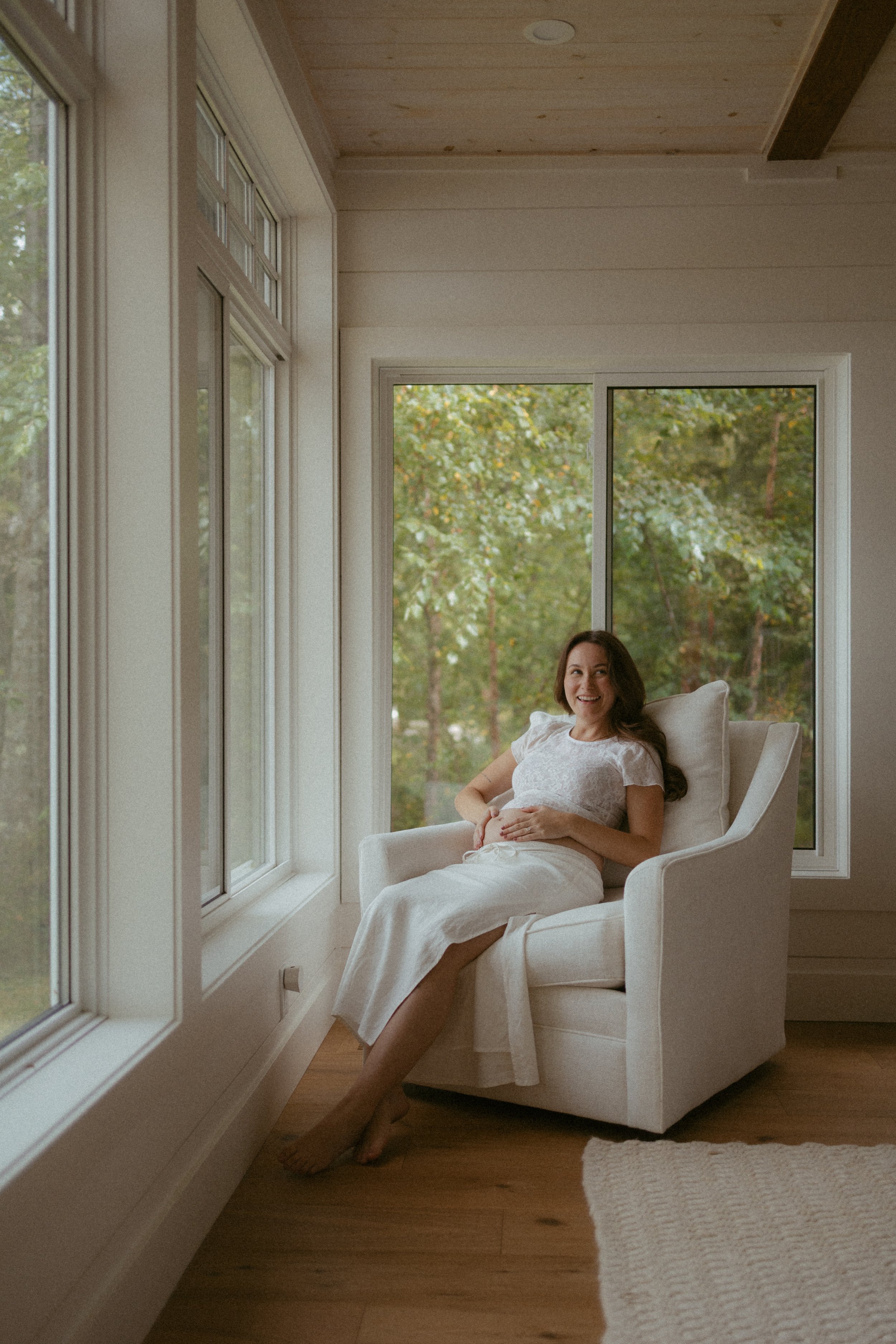 A woman sitting in a white armchair in a bright room with large windows and a view of greenery outside. She is smiling, wearing a white dress, and appears to be pregnant, with her hands resting on her belly.