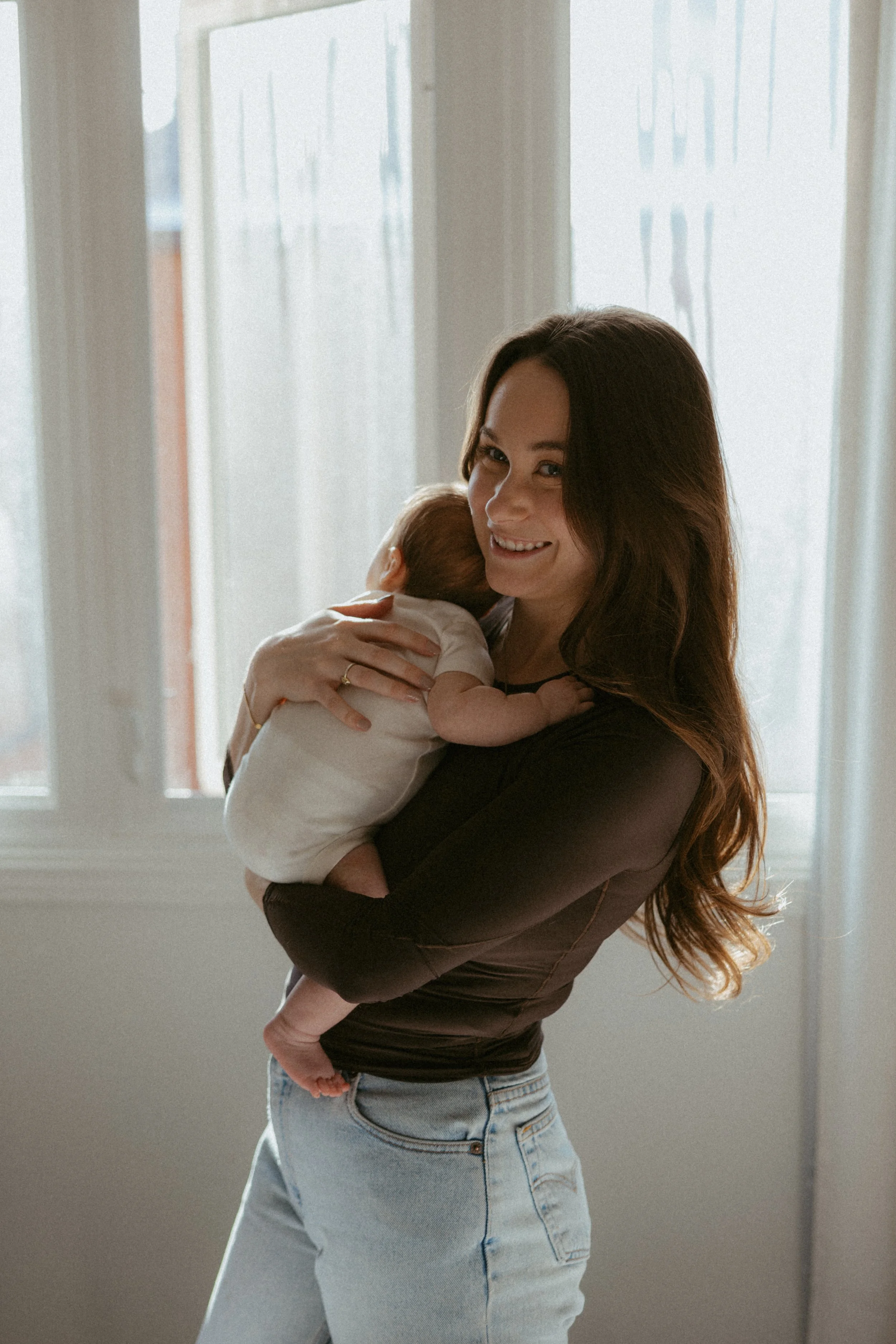A woman with long brown hair, wearing a black top and light blue jeans, smiling while holding a baby in front of a window with sheer curtains.
