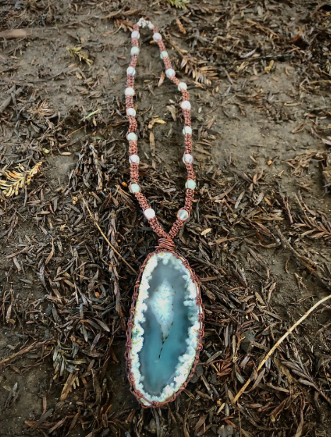 A necklace with a large blue and white geode pendant hanging on dirt with dried leaves.
