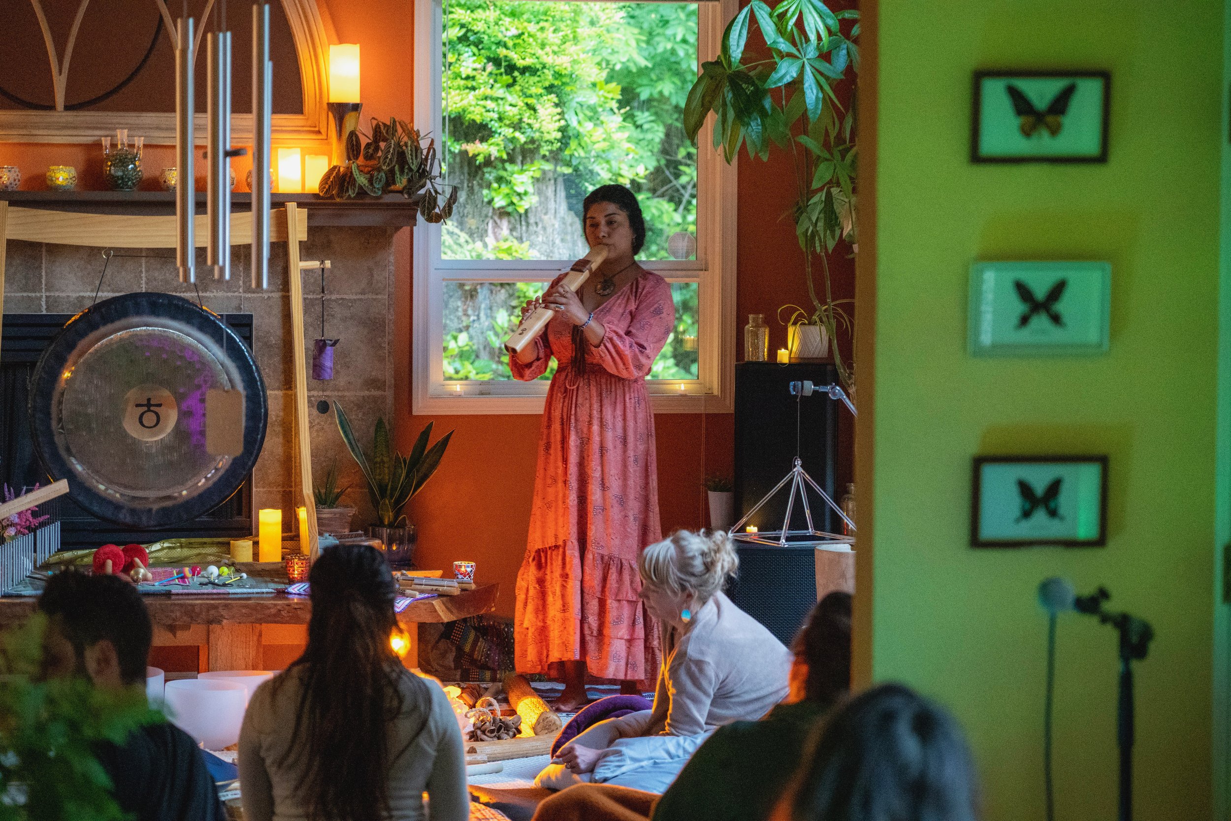 A woman in a pink dress playing a wooden flute in a room with orange and green walls, with children sitting on the floor in front of her, some looking at her and some facing away. The room is decorated with plants, candles, and butterfly pictures on the walls, and a window showing trees outside.