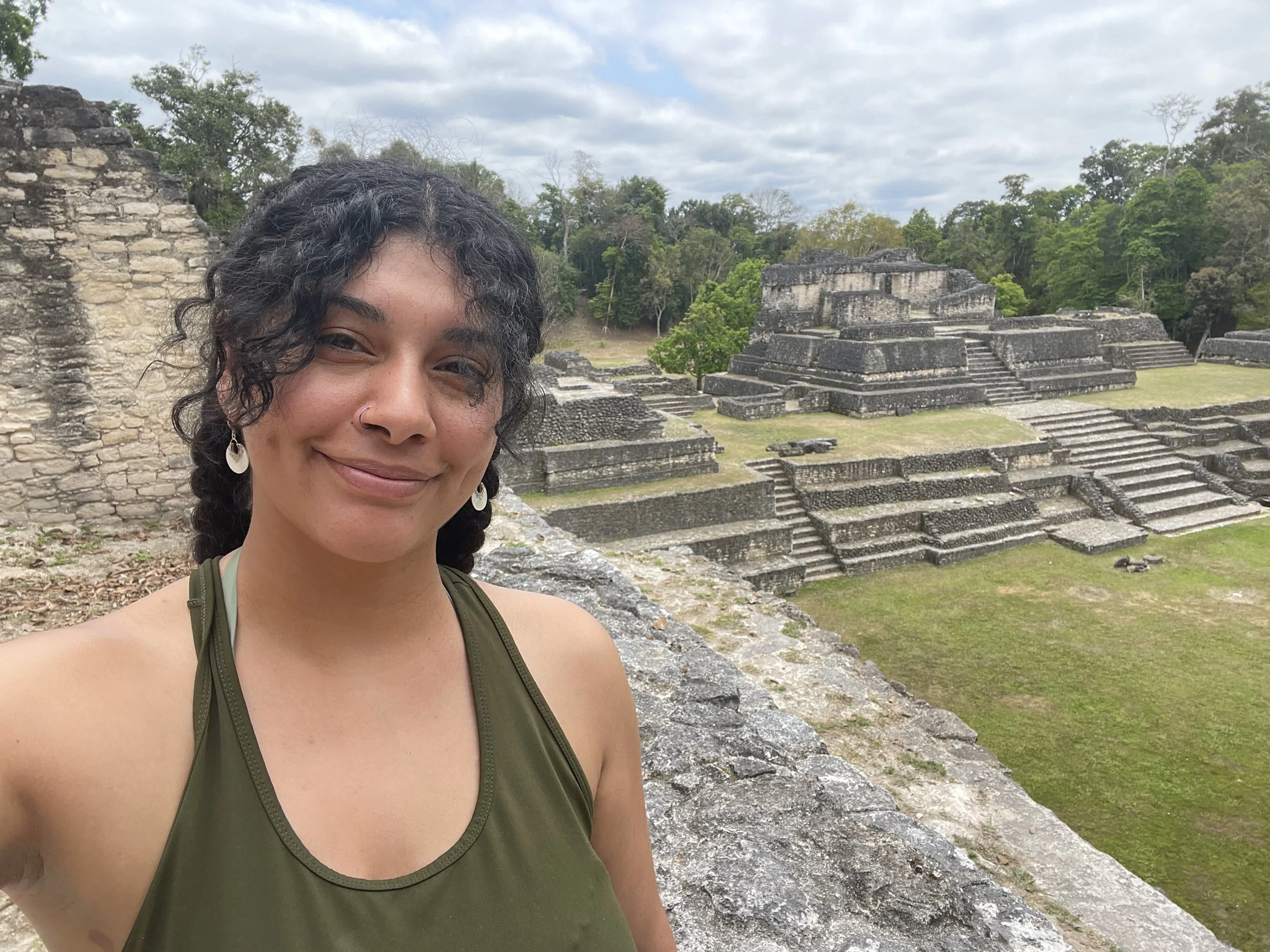 A woman taking a selfie at an ancient Mayan ruin site surrounded by greenery under a cloudy sky.